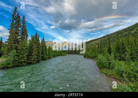 Scenic view of a rainbow in the blue sky against the clouds above Grey Ridge as the Wheaton River flows below towards them; Whitehorse, Yukon, Canada Stock Photo