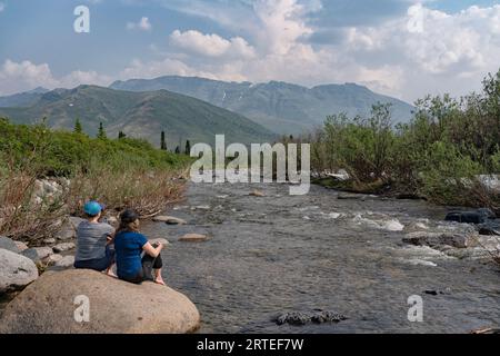 View taken from behind of two women, friends sitting on a rock and watching the Klondike River blow by during the hot days of the Yukon summer Stock Photo