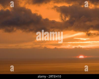 Dramatic sunset through clouds and over the ocean's horizon, showing a warm glow of orange over the sky and water; Hawaii, United States of America Stock Photo