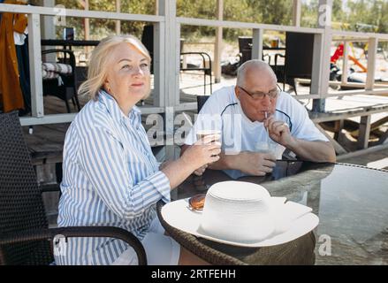An elderly couple is sitting at a table in a cafe by the sea, looking at the sea, drinking a cocktail and coffee. Stock Photo
