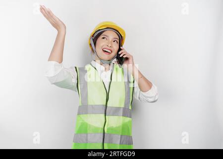 A smiling Asian woman labor wearing safety helmet and vest, talking on her phone and pointing to copy space above her, isolated by white background. L Stock Photo