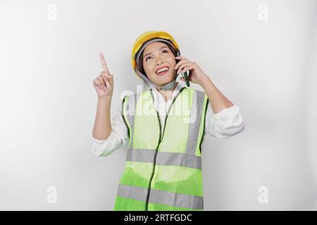 A smiling Asian woman labor wearing safety helmet and vest, talking on her phone and pointing to copy space above her, isolated by white background. L Stock Photo