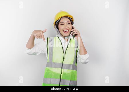 A smiling Asian woman labor wearing safety helmet and vest, talking on her phone and pointing to copy space below her, isolated by white background. L Stock Photo
