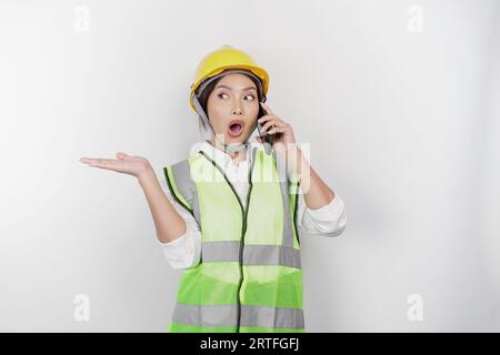A smiling Asian woman labor wearing safety helmet and vest, pointing to copy space beside her while having a phone call, isolated by white background. Stock Photo