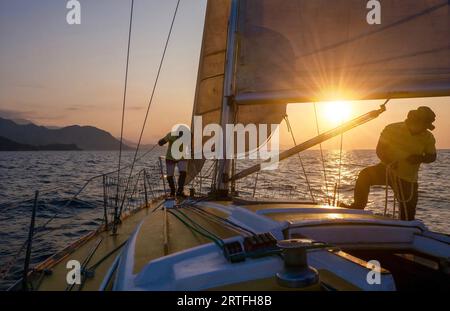 Puerto Galera, Philippines - March 21, 2023. A small recreational sailboat at sunset, with two male crew members making sail changes. Stock Photo