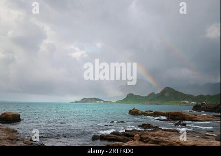 There are two rainbows in the sky by the sea. Rocky coast under blue sky and white clouds. White waves and light blue sea. Stock Photo