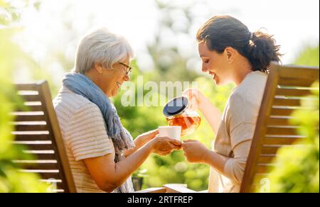 Happy young woman and her mother drinking tea in summer morning. Family sitting in the garden with cups and enjoying the conversation. Stock Photo