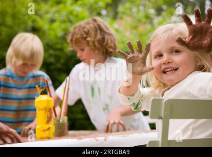 Young girl sitting at table holding arms up with hands covered in paint Stock Photo
