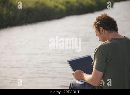 Back view of a man sitting by a river using a laptop computer Stock Photo