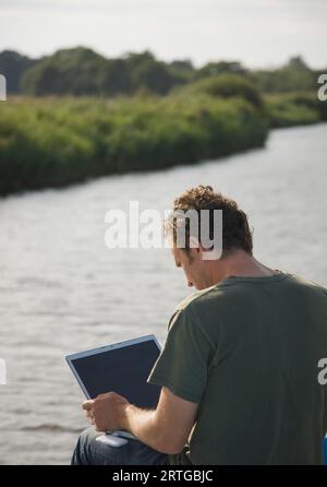 Back view of a man sitting by a river using a laptop computer Stock Photo