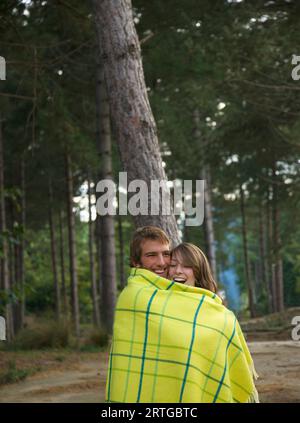 Young couple standing in a forest wrapped in a blanket laughing Stock Photo