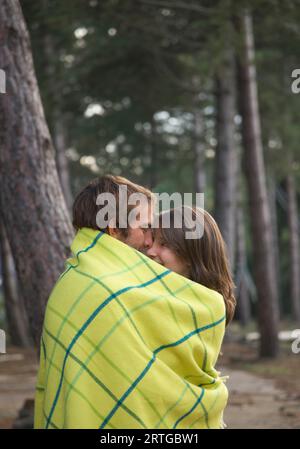 Young couple standing in a forest wrapped in a blanket looking at each other Stock Photo
