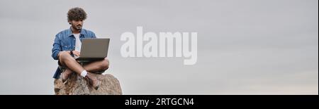 digital nomad, focused indian man using laptop, sitting on rock, remote work concept, banner Stock Photo