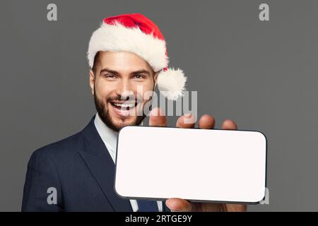 Portrait of happy handsome businessman wearing red Santa hat holding smartphone with blank empty white screen display, Christmas concept Stock Photo