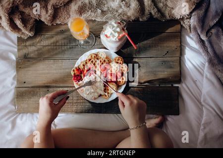 Young woman eating appetizing waffles and tasty drinks on tray on bed Stock Photo