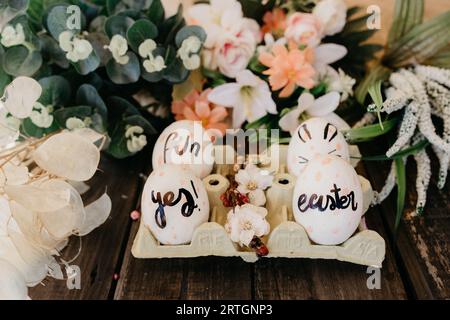 Closeup set of colored chicken eggs in container between ornamental flowers on wooden background Stock Photo
