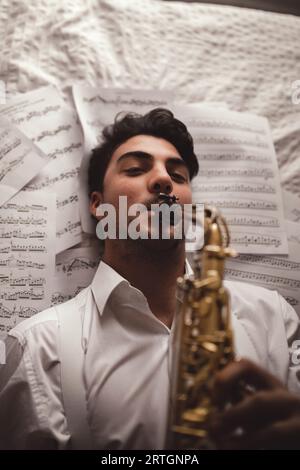 From above young romantic musician playing on saxophone and lying on sheets of printed music Stock Photo