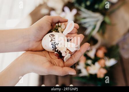From above crop hands of female holding colored chicken egg with decorative rabbit ears and flowers on blurred background Stock Photo