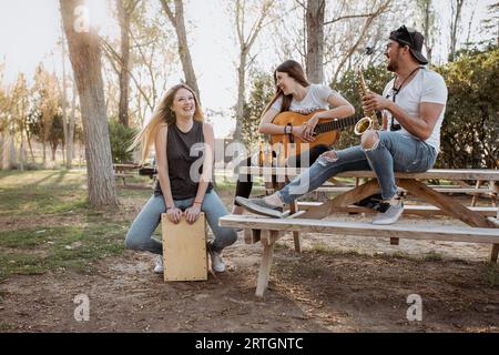 Young man and women with musical instruments sitting near table and laughing on sunny day in park Stock Photo