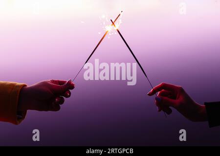 Crop female hands in front of each other holding burning sparklers on colorful purple background Stock Photo