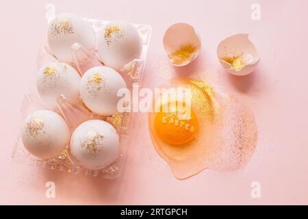 Top view of broken glittered egg with text Happy Easter and raw eggs in plastic box on pink background Stock Photo