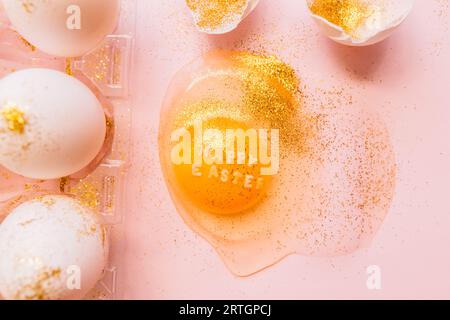 Top view of broken glittered egg with text Happy Easter and raw eggs in plastic box on pink background Stock Photo