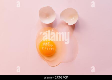 From above raw broken egg with text Happy Easter on yellow yolk and eggshells on pink background Stock Photo