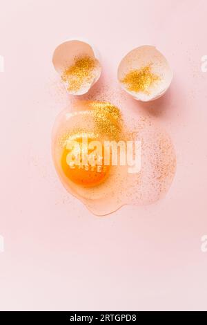 From above raw broken egg with text Happy Easter on yellow yolk covered in glitter and broken eggshells on pink background Stock Photo