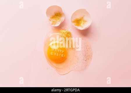From above raw broken egg with text Happy Easter on yellow yolk covered in glitter and broken eggshells on pink background Stock Photo