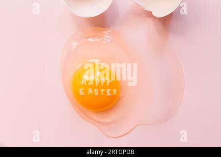 From above raw broken egg with text Happy Easter on yellow yolk and eggshells on pink background Stock Photo