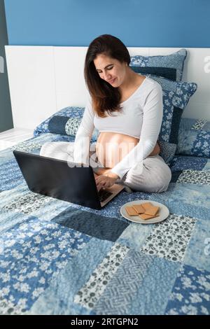 From above of smiling pregnant female browsing netbook while sitting on comfortable bed at home Stock Photo