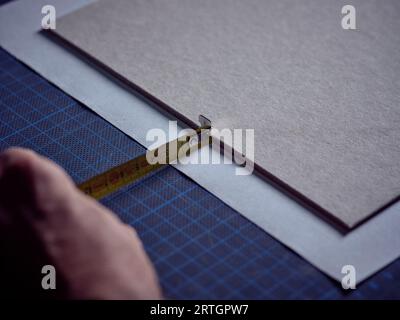 Unrecognizable crop male artisan using tape and measuring paper on blue mat while working in bookbinding workshop Stock Photo