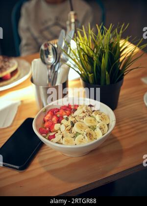 From above bowl of delicious breakfast topped with fresh strawberries and bananas served on table with potted plant in light cafe Stock Photo