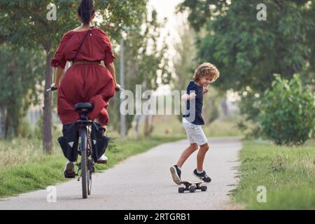 Back view of young woman riding bike and boy near her riding skateboard performing cross feet tricks Stock Photo