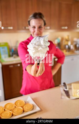 Happy blur female pastry chef smiling and looking at camera while showing cream on mixer near table in kitchen Stock Photo