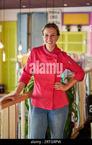 Smiling confident female chef in professional clothes with hand on waist standing near wooden railing in colorful room and looking at camera Stock Photo