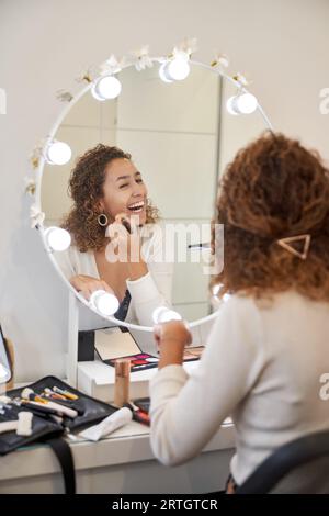 Back view of happy black female with curly hair applying foundation with brush while laughing and looking at mirror Stock Photo