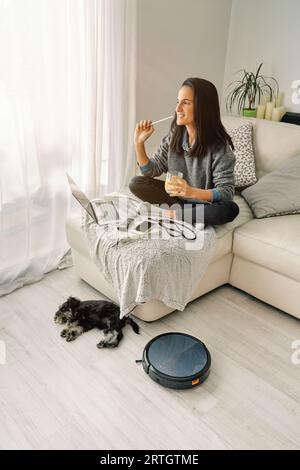 Side view of female with brown hair sitting with glass of drink on sofa with opened laptop and smartphone in room with sleeping dog and robotic vacuum Stock Photo