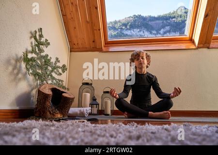 Full body of barefoot boy meditating with closed eyes while sitting in Padmasana on yoga mat at home Stock Photo