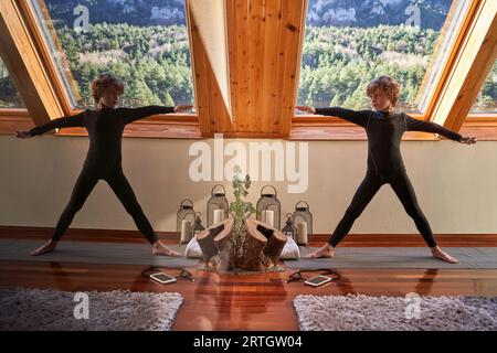 Full body of slim boy in black activewear doing exercise while practicing yoga on mat in spacious apartment and looking at mirror Stock Photo