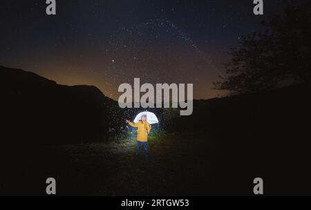 Unrecognizable traveler in yellow jacket standing alone among dark trees with raised arm and umbrella at rainy night Stock Photo