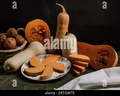 Composition of potatoes in wicker basket halved butternut squash with seeds and slice nutmeg pumpkin in plate placed on table with white cloth against Stock Photo