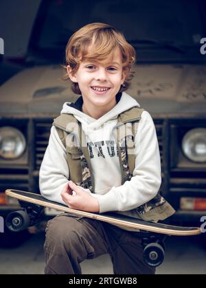 Happy boy in stylish outfit holding skateboard and standing near car on street while smiling and looking at camera Stock Photo