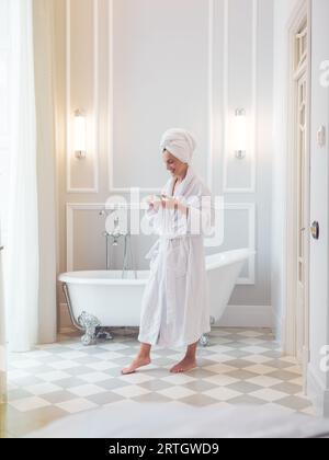 Side view of young barefoot female wearing white bathrobe and towel on head standing near bathtub with cup of coffee in hotel bathroom at morning time Stock Photo