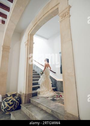 Back view of full body young female newlywed wearing elegant bridal dress and with bunch of flowers looking up while walking upstairs holding on banis Stock Photo