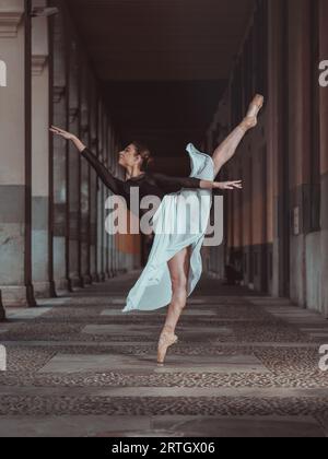 Full body of graceful ballerina in silk skirt and pointe shoes balancing in arabesque ballet position in empty passage of building Stock Photo