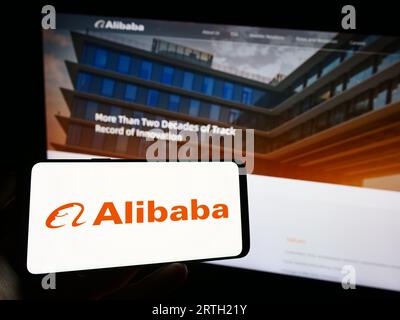 Person holding cellphone with logo of Chinese company Alibaba Group Holding Limited on screen in front of webpage. Focus on phone display. Stock Photo