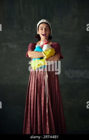 Happy young woman in costume of medieval maid standing with laundry against vintage green background Stock Photo