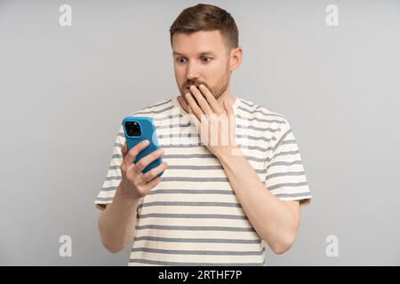 Shocked scared man closing mouth with hand looking at smartphone screen on grey background. Stock Photo