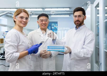 Portrait of an international group of chemists, doctors, scientists, men and women standing in a laboratory in uniforms and holding flasks with samples for research. They look and smile at the camera. Stock Photo